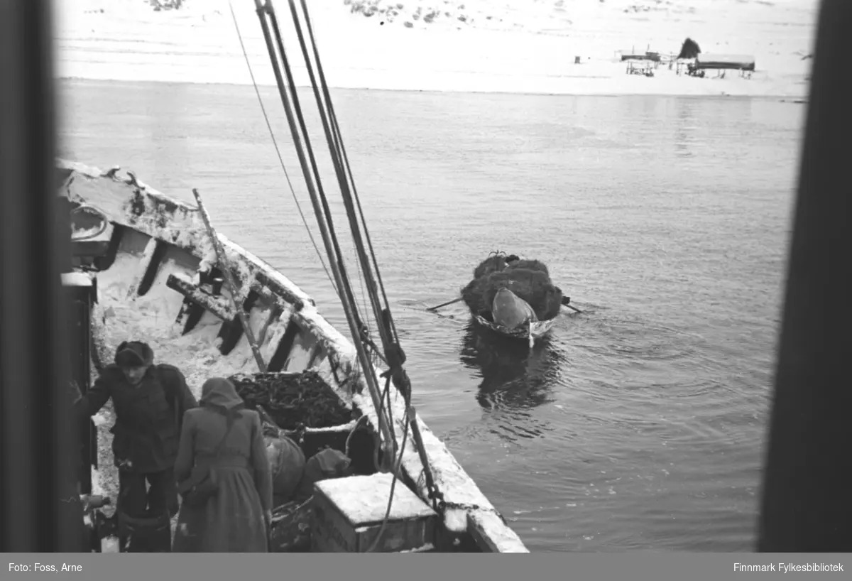 Arkitekt Arne Foss har fotografert dette bildet fra båten mellom Billefjord-Honningsvåg i februar-mars i 1947. Gjenreisning er i gang i Finnmark. En robåt nærmer seg kaia med en stor høylass.