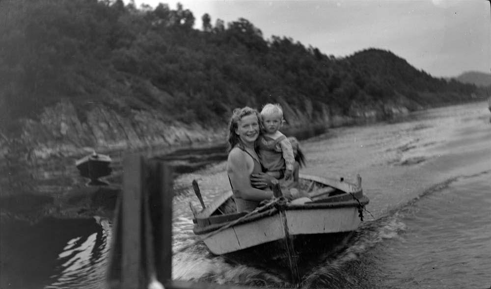Jente som holder et barn, sitter i robåt som blir trukket av større båt.