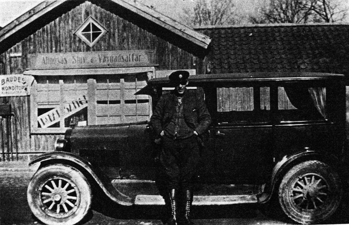En man står med händerna i fickan och lutar sig mot en personbil. Bakom honom syns skyltar med text "Baudes Konditori" och "Alingsås Stuv & Vävnadsaffär"