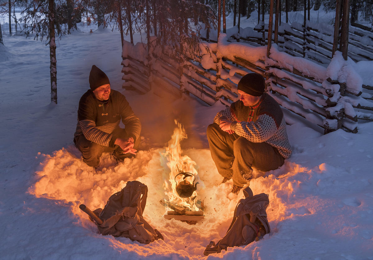 To personer i ullgenser og vadmelbukse foran et bål på en vinterkledd Prestøya