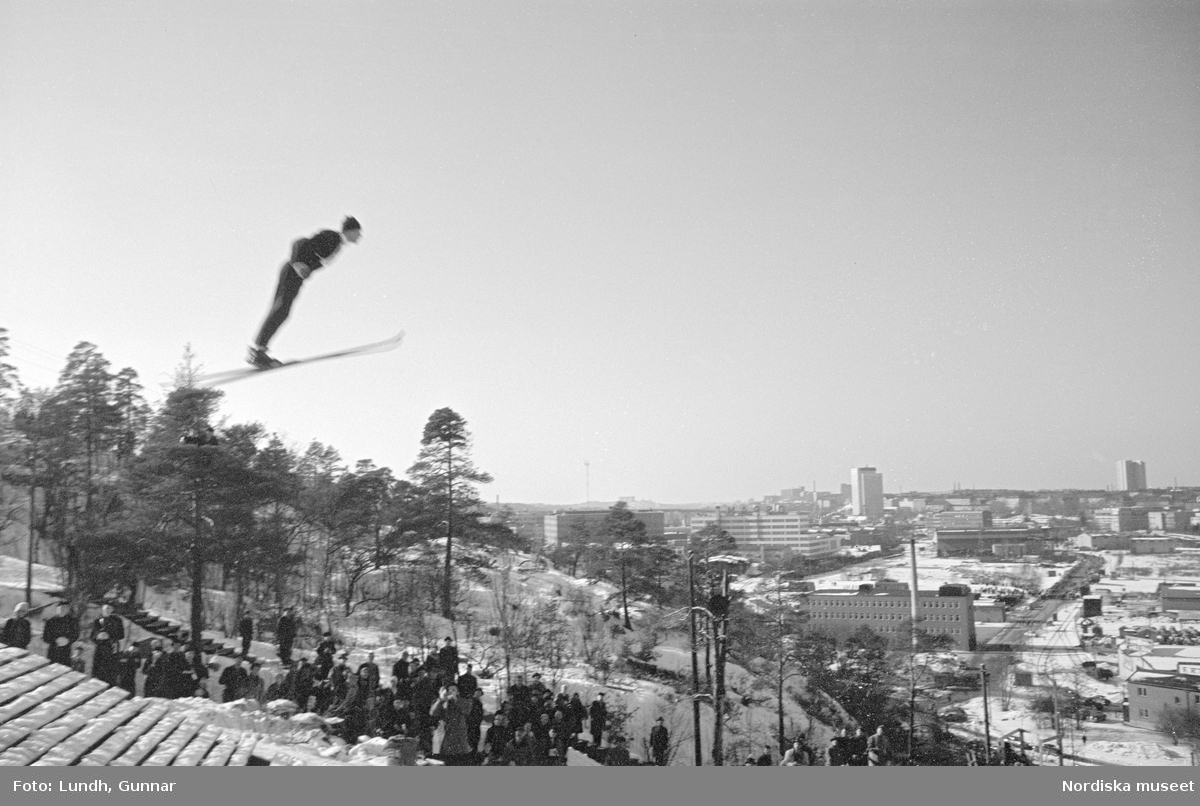 En tävling i backhoppning i Hammabybacken, Stockholm, backhoppare hoppar från en hoppbacke inför åskådare och domare, vy över Södra Hammabyhamnen och Södermalm.