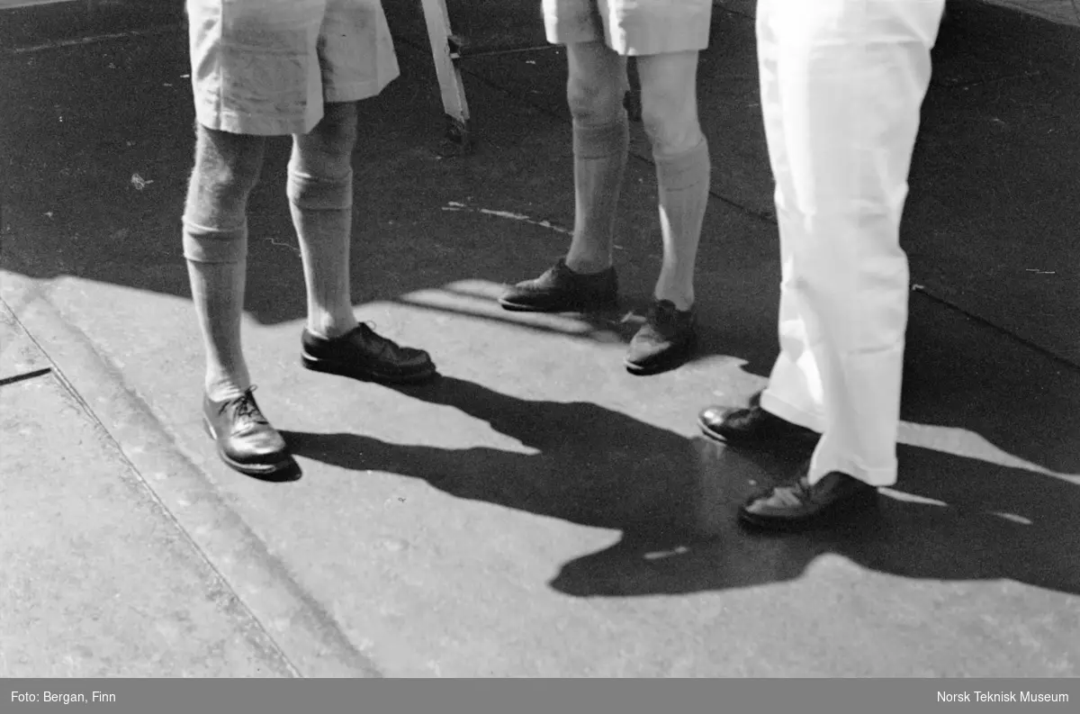 Føtter, ben til tre personer som står på et skipsdekk, sol og skygger