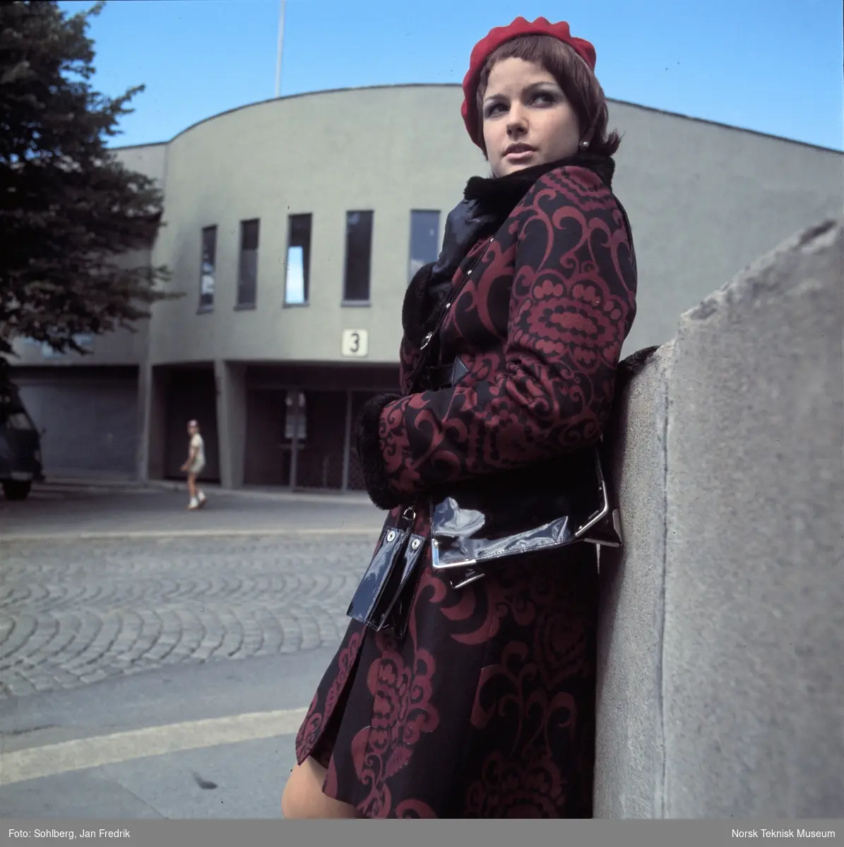 Motebilde, kvinne i mønstrete rød kåpe med alpelue, foran idrettsarena. Bildet er en del av en serie der noen er publisert i ukebladet Det Nye 1969, nr. 38.