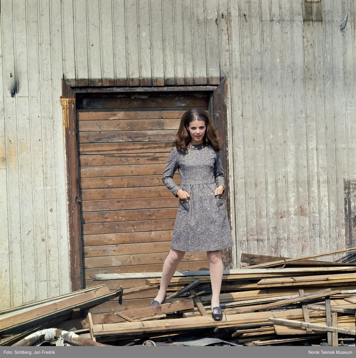Modell kledd i mønstrete kjole foran et falleferdig hus.