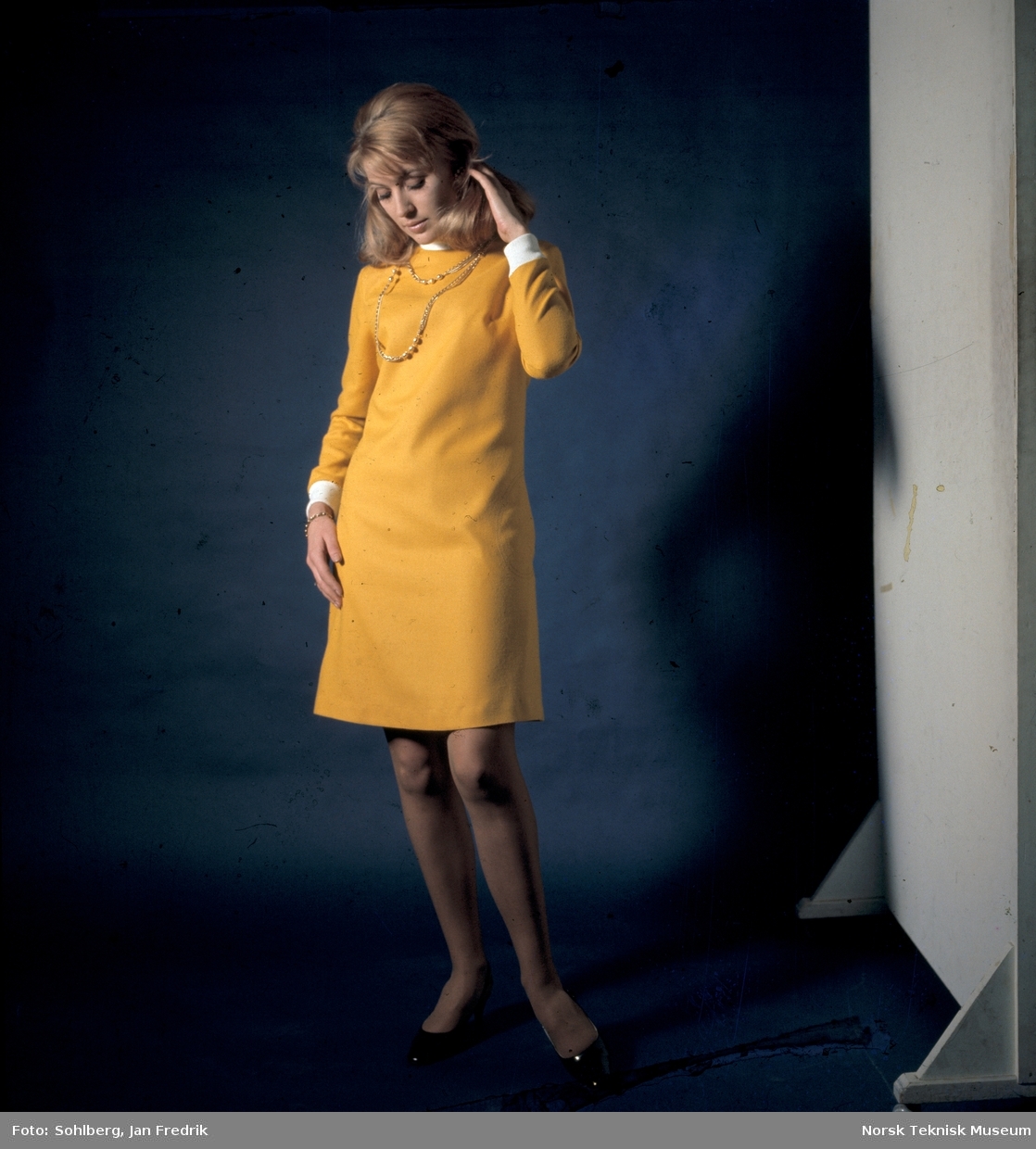 Tidlig norsk motefotografi. Kvinnelig modell poserer i en gul kjole mot blå bakgrunn.