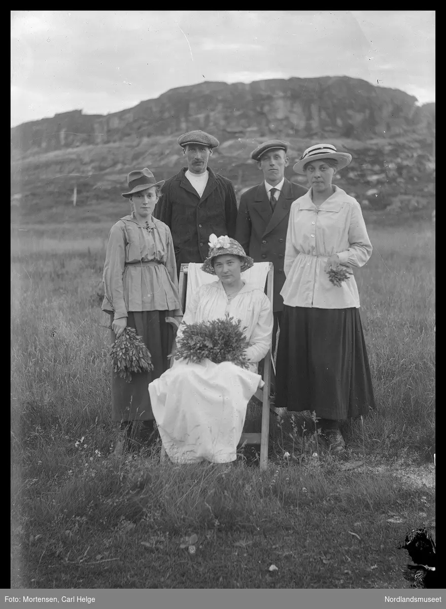 To unge menn og tre unge kvinner/jenter, hvorav ei i hvitt kjole på en stol. De andre står rundt henne på en eng, i bakgrunnen en bergknaus.