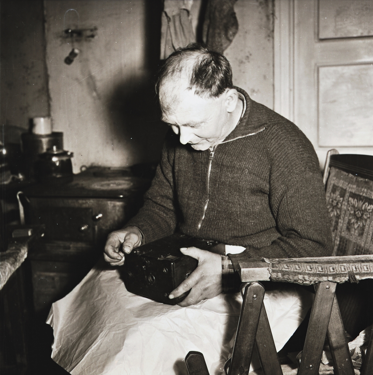 Bygdefotografen "Kloar-Erik" sitter vid spisen i sin stuga med en av sina äldsta lådkameror. Bilden publicerades i Varbergsposten 1953.