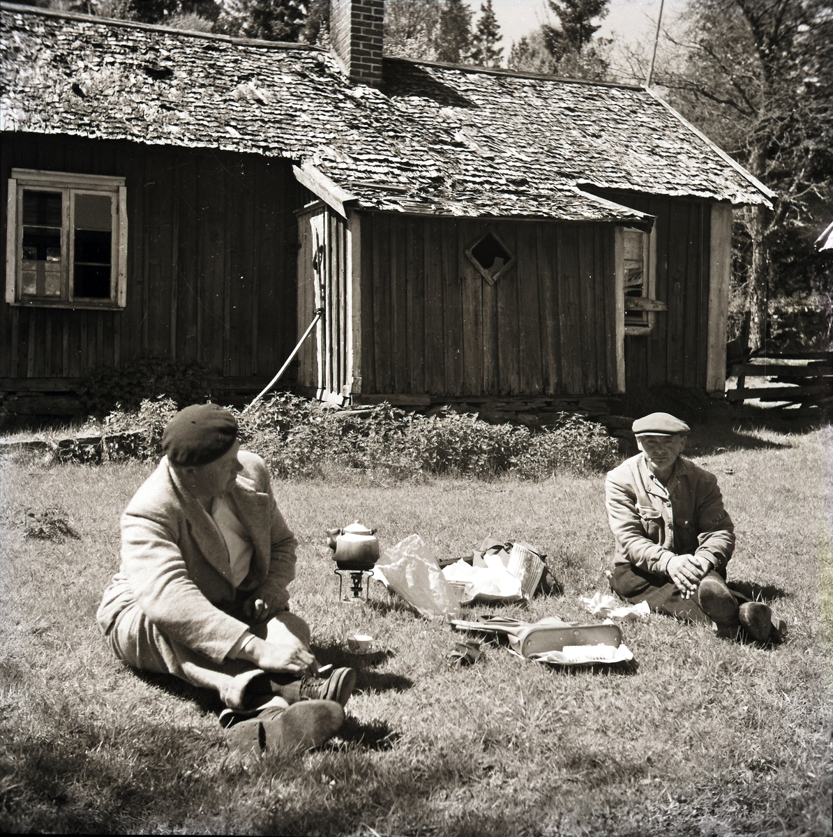 Bygdefotografen "Kloar-Erik" och Anders Lundgren sitter i gräset vid en stugan i Dämmarehult och dricker kaffe som de har kokt i kaffepanna på ett spritkök. Anders röker en cigarett.