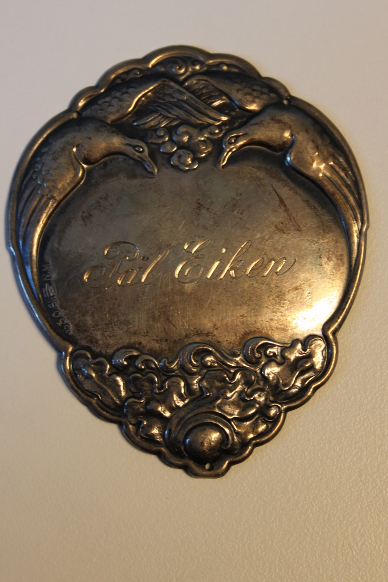 Sølvfarget plakett inngravert med navnet "Pål Eiken". Utsmykket med to duer, utskjæringer og navnet i midten.