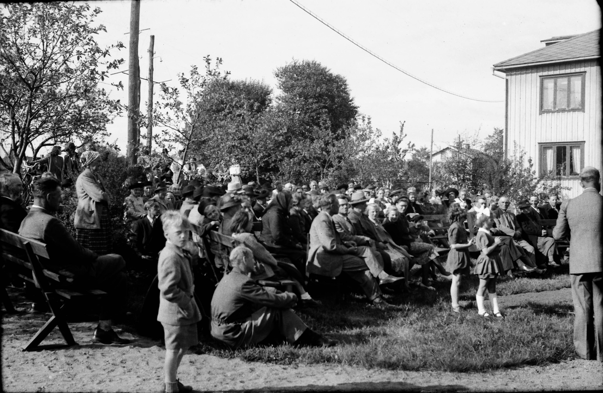 Friluftsmöte med Betaniaförsamlingen i Veddige. En stor skara människor sitter på träbänkar i en trädgård.
(Se även bildnr EA0972)