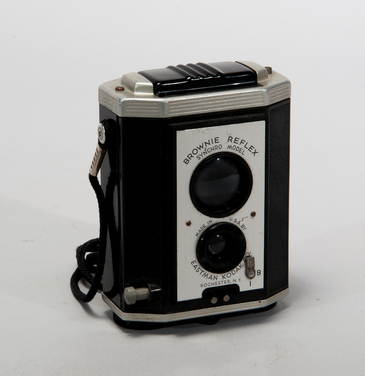 Tvåögd spegelreflexkamera för mellanformat, tillverkad i bakelit och metall. Med axelrem i textilmaterial.
Märkt: Brownie Reflex Synchro Model.