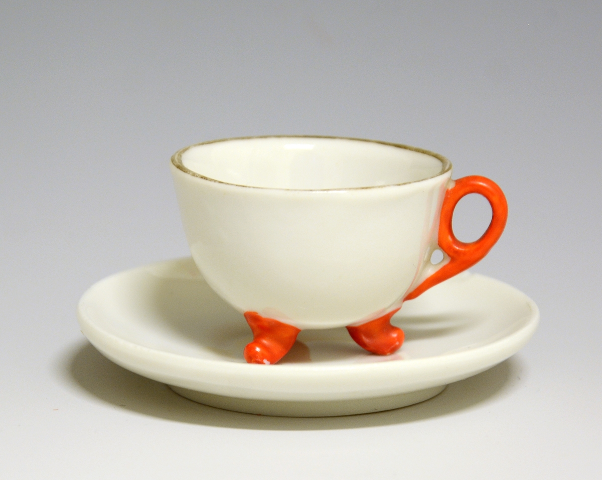 Kopp og skål til dukkeservise i fajanse, hvit glasur med gulldekorerte kantrender. Koppen har rød hank og står på tre føtter som også er malt røde.