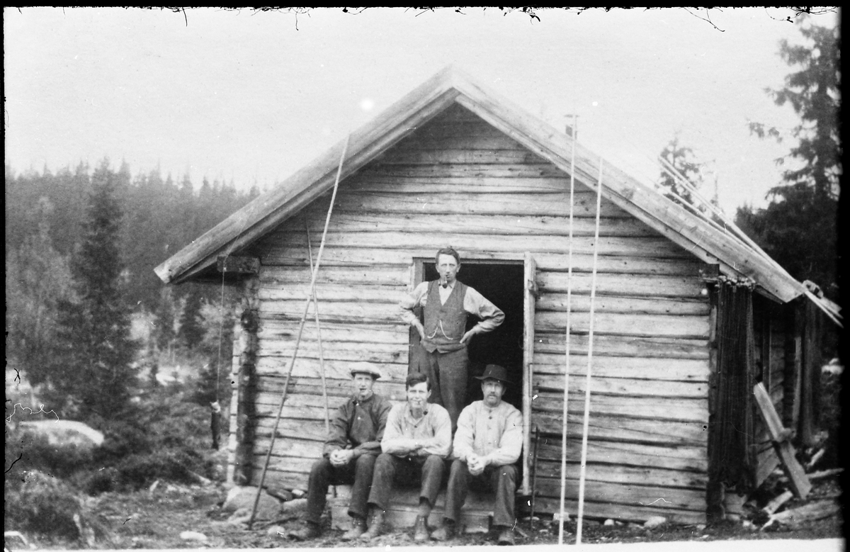 Avfotografert bilde av fire menn foran ei tømret skogshytte. Fiskestenger står reist opp mot veggen. Hverken personer eller sted er identifisert.