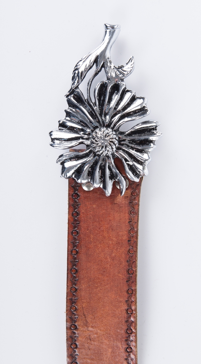 Lærbelte med antakelig svistempet dekor. Metallspenne formet som  blomst med stilk og bLad. Lærhempe til å stikke enden inn i for ekstra feste (FHM.09272.b)
Kjøpt som souvenir på Mallorca 1977