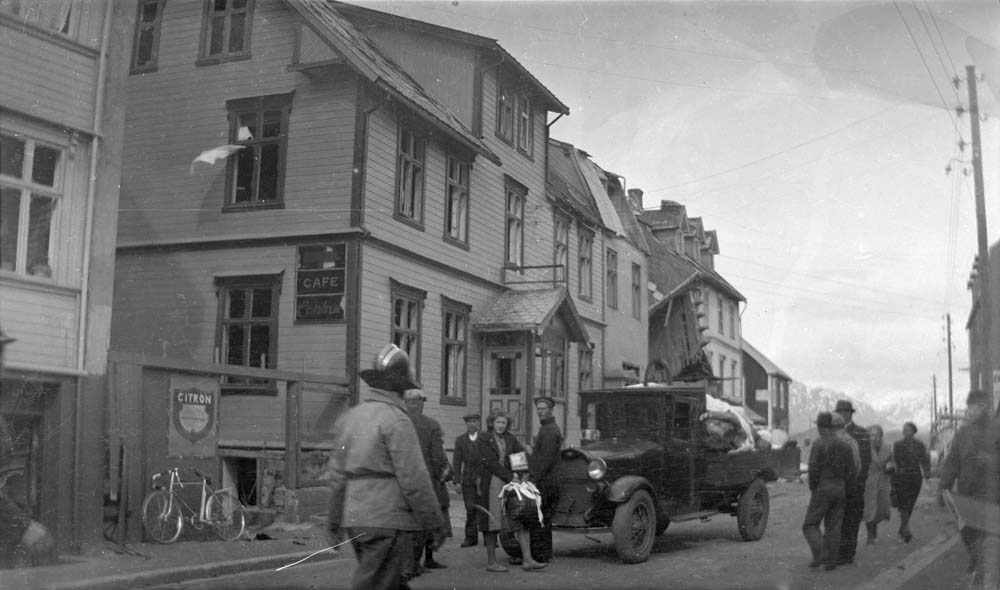 Cafe Central i Sandnessjøen med Hotell Bristol bortenfor. Brannmann, flere personer i gaten. Kanskje fra da Hotell Bristol ble bombet i 1940. Lastebilen er en Ford AA 1928-29-modell.