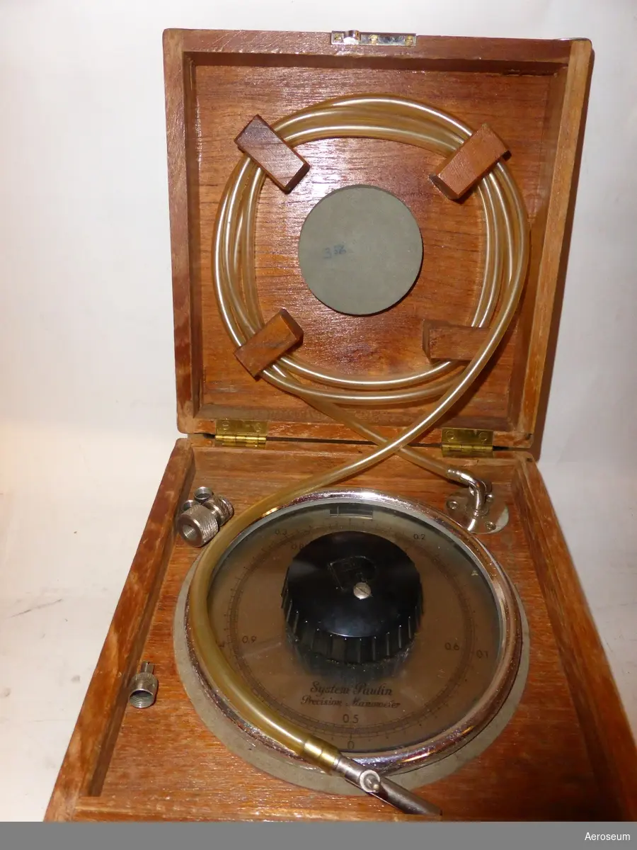 En precisionsmanometer i lackat trähölje, tillverkad av L M Ericsson, med manometer från System Paulin. I lådan sitter en display med en vridbar ratt och nålvisare och en genomskilig slang som sitter fast i ena enden med en metallbit i andra.