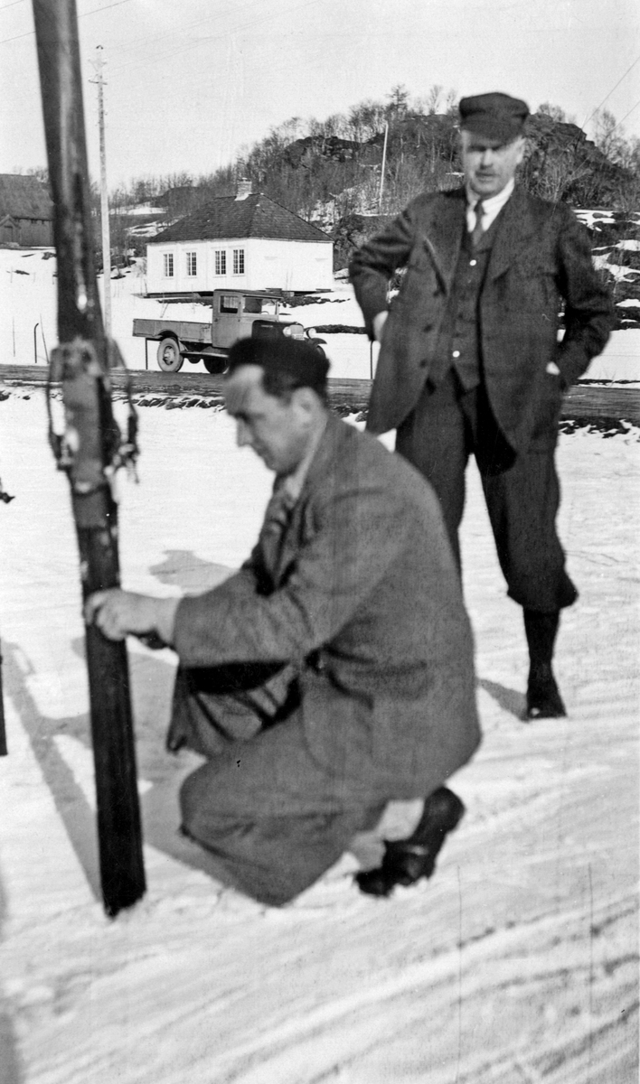 Vinterbilde med Trøndelag folkemuseum i bakgrunnen. Stående ses Christian Anker Bachke.
"Mars 1934" påskrevet på baksiden.