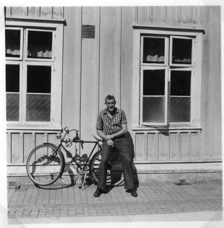 En man i rutig skjorta, mörka byxor och cigarett i handen sitter på pakethållaren till en cykel mot en husvägg, möjligen Hotell Brahe.