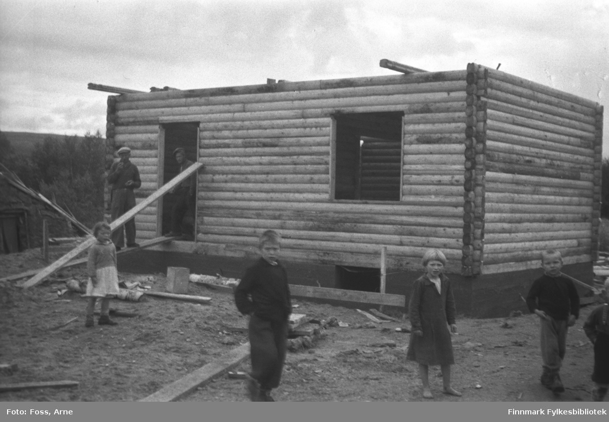 Et tømmerhus under bygging i Tana, i mai-juni 1947. Fem barn i forgrunnen og to menn ved døråpningen.