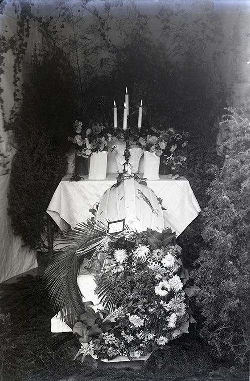En ljus likkista står på en avsats täckt med ett ljust lakan, framför ett bord med duk, fem ljus och blommor. En krans vid kistans fotände.