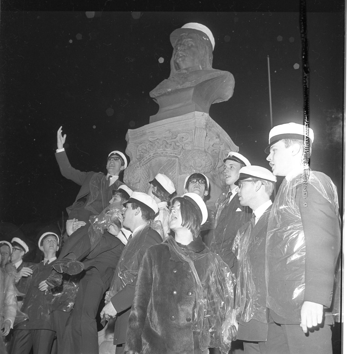Studenter vid Grännaskolan, 1967, samlade under Brahebysten på Gränna torg. De vinkar och sjunger, troligen. De har transparent plastskydd över kläderna. Brahe har fått en studentmössa på huvudet.