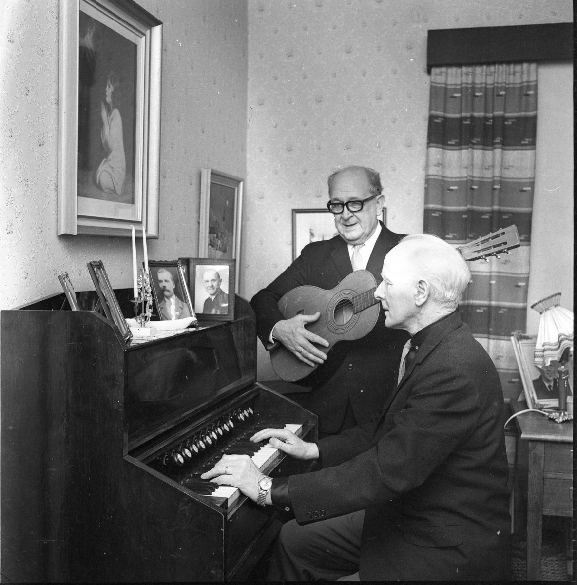 Carl Öst i glasögon spelar på en gitarr. Han står intill Martin Persson som sitter och spelar på en orgel. På orgeln står en ljusstake med två ljus samt ramade porträttfotografier av män.
