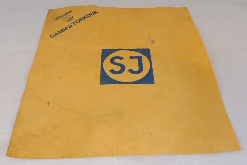 Rektangulär, beige-gul duk. Högst upp i höger hörn finns texten: "Vileda DAMM & TORKDUK", tryckt i blått. På mitten finn: "SJ", samt SJ:s logotyp tryckt i blått.