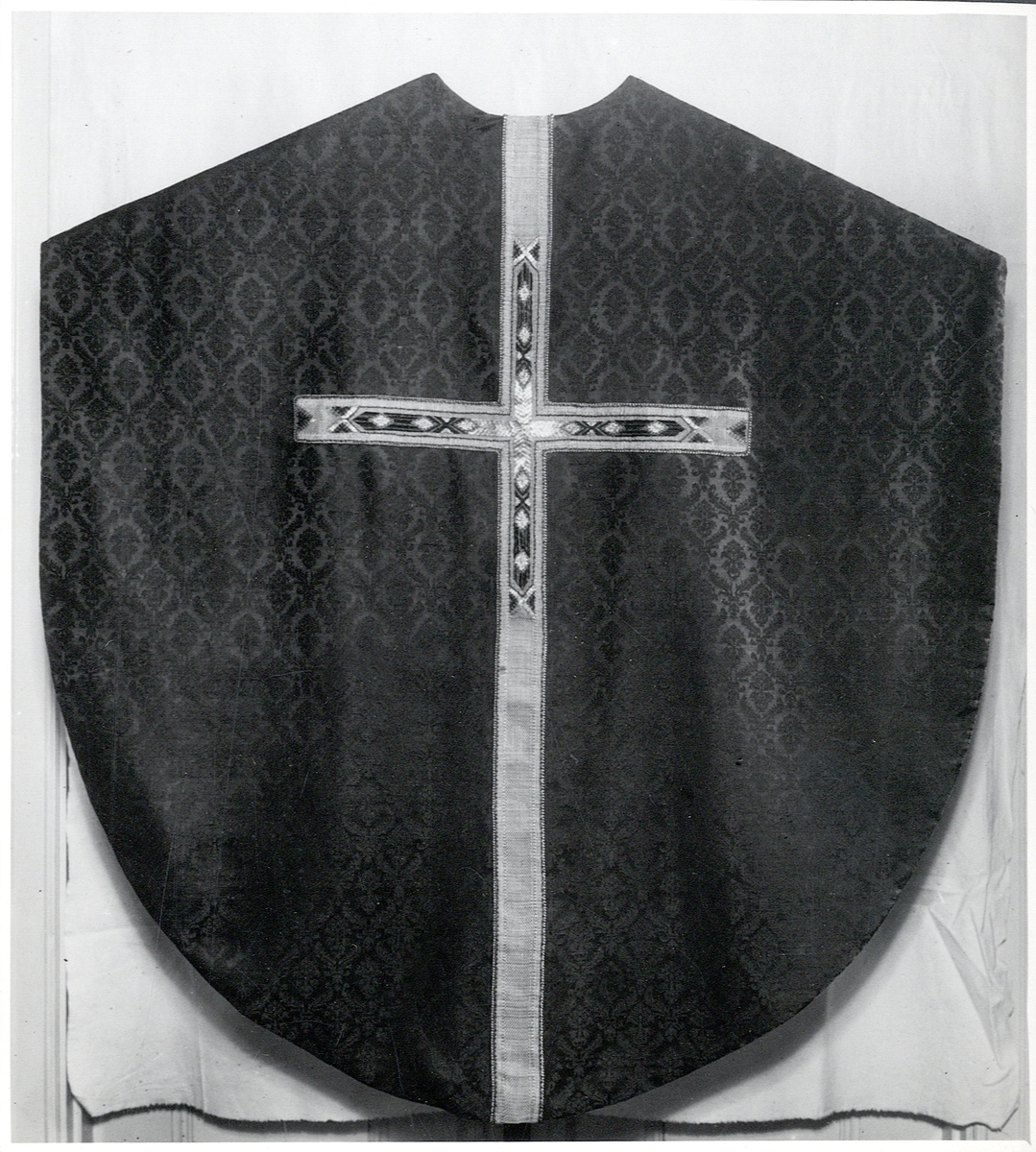 Foto (svart/vitt) av en mörk mässhake (baksidan) i brokad med ljusare, broderat korsparti.

Inskrivet i huvudbok 1983.