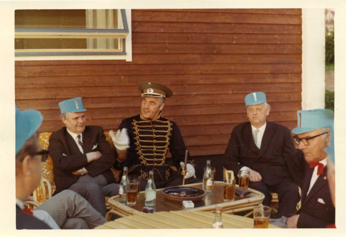 Minst sju män sitter utomhus runt ett bord med flaskor och glas med okänt innehåll, på bordet finns också en askkopp och ett paket cigaretter. Männen har kostym med slips eller fluga och på huvudet en blå hög skärmmössa. Mannen i mitten avvikter med en Dolma m/1895 och på huvudet en militär keps av modernare snitt. De sitter vid en rödfärgad byggnad med panel på förvandring. Från vänster: Hans Persson, Holger Andersson, Olle Mathias, Sture Petri och Osse Gustafsson.