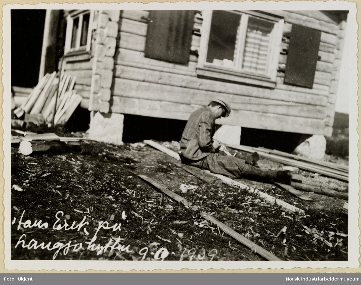 Hansk Erik sitter på tømmer foran laftet hytte og arbeider