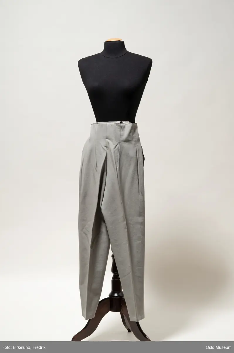 Lange grå bukser i elastisk, syntetisk stoff. Lukkes med glidelås i siden.
