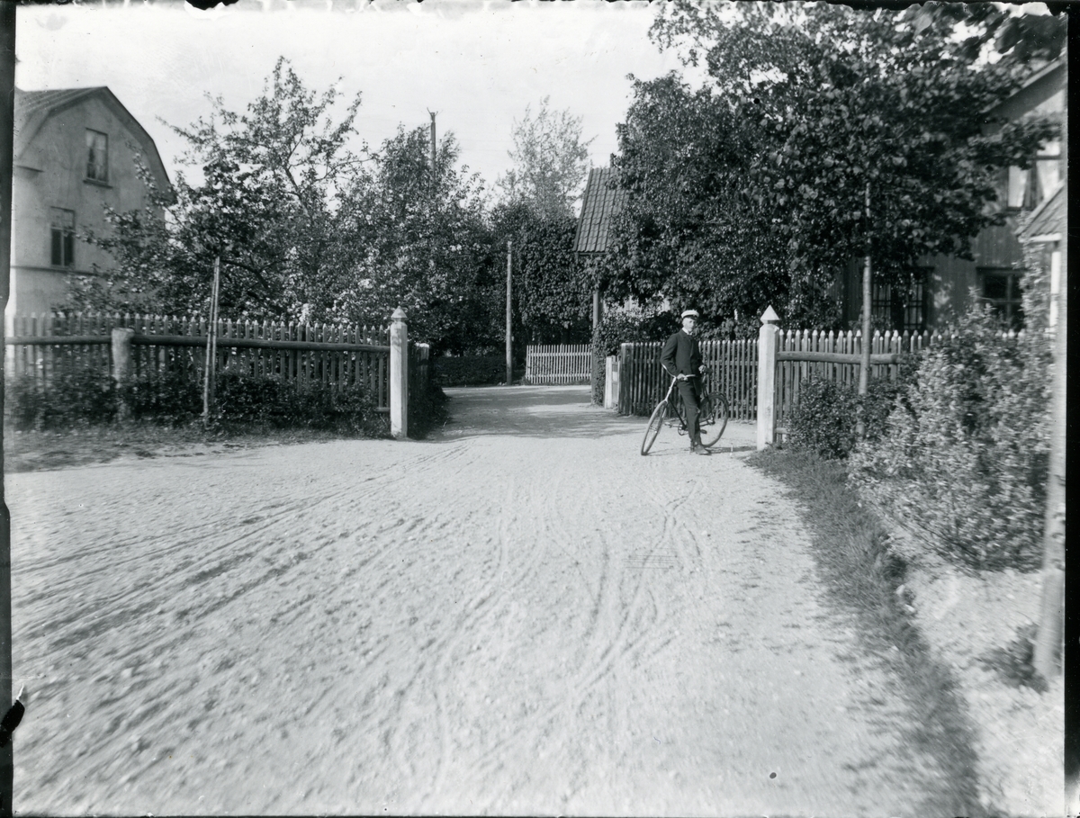 Kung Karl sn, Kungsör.
Pojke (ung man) står vid cykel på en grusplan, med staket  och villor i bakgrunden. 1913.
Text under bilden "Helleday från folkskolan"