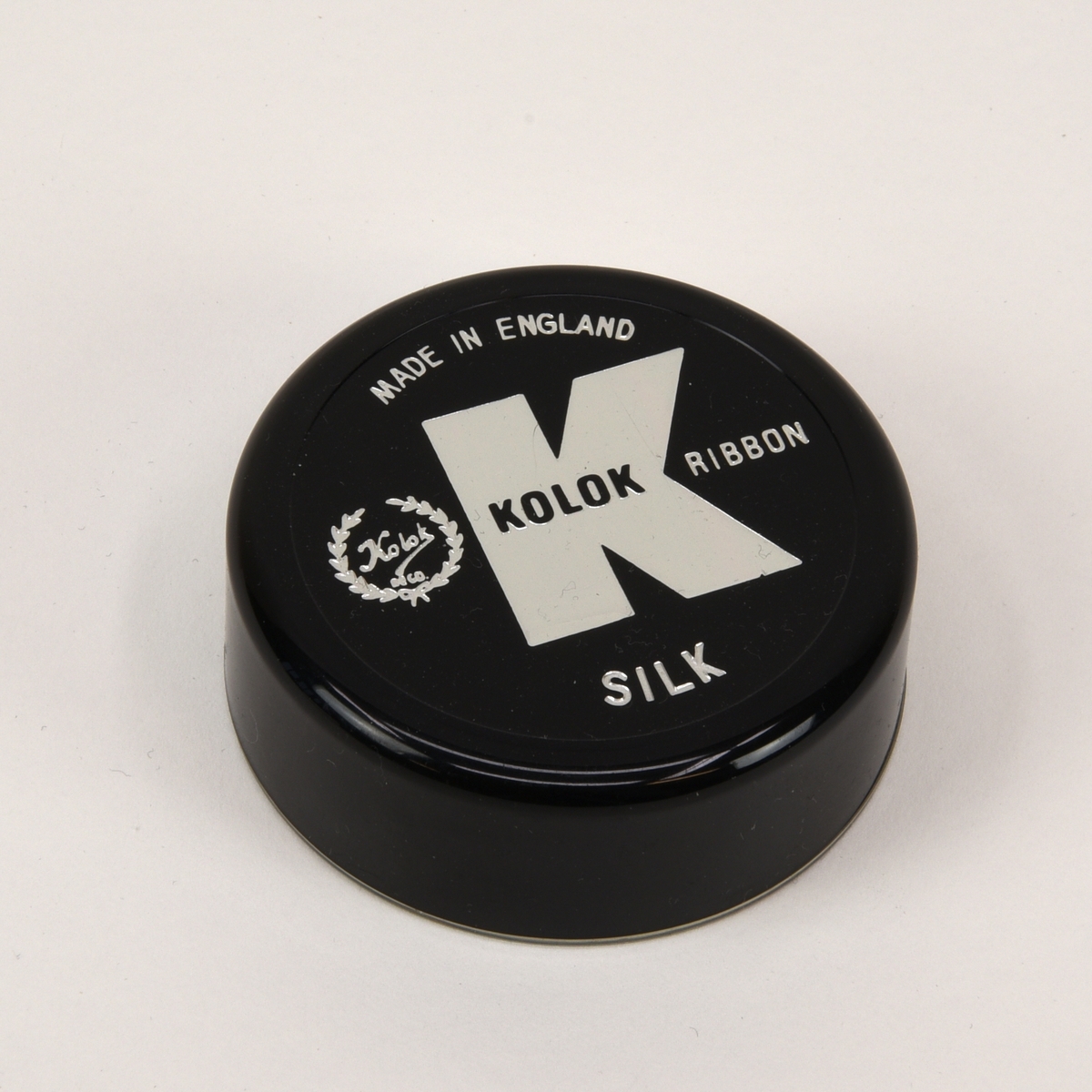 Två svarta 13 mm färgband till skrivmaskin på spolar av DIN 2103-standard. Båda band ligger i varsin plastask med lock.

Det ena färgbandet (:18) är använt och upprullat på en spole från Pelikan men ligger i en rund ask från Kolok (:19-20). Asken har en genomskinlig underdel och ett svart lock. På locket finns Koloks logotyp, "Made in england" och "Silk" tryckt i silver. I botten av asken finns ett guldfärgat papper med information om vilka skrivmaskinstyper bandet passar till.

Det andra bandet (:21) är oanvänt men saknar helt information om tillverkare och dylikt. Det enda som finns är "DIN 2103" instansat på spolen och ett eventuellt artikelnummer på askens undersida. Spolen är förpackad i folie och asken (:22-23) är av samma typ som (:19) men locket är rött och saknar text.