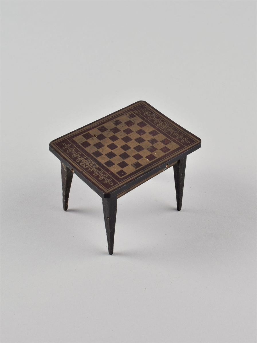 Lite rektangulært bord med bred, dekormalt sarg og fire rette ben. Påmalt gullfarvet sjakkbrett og blomsterranker på platen.