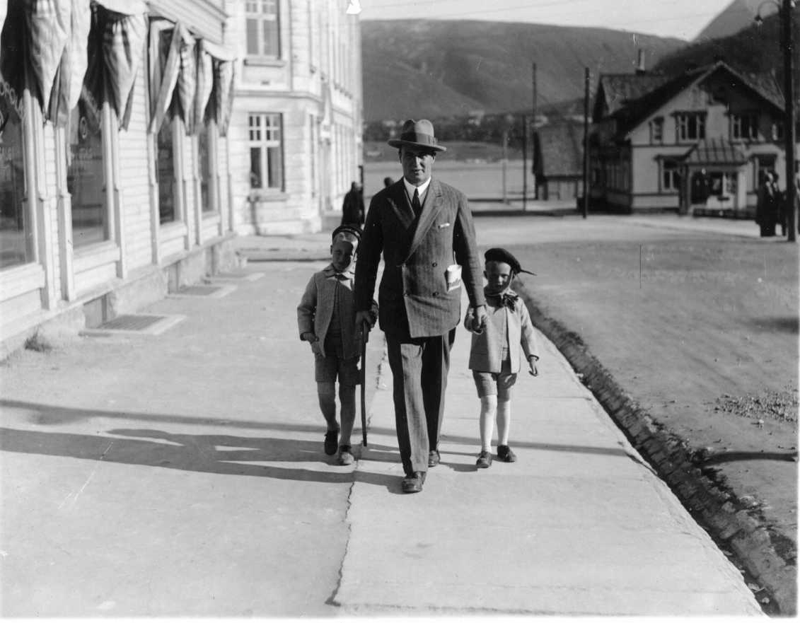 En man i kritstrecksrandig dubbelknäppt kostym med slips och hatt promenerar sannolikt i Tromsö. I fickan har han en vikt tidning, Aftenposten. Han håller ett barn i vardera hand. Barnen har höga vit strumpor, knäkorta byxor med jacka i samma tyg.