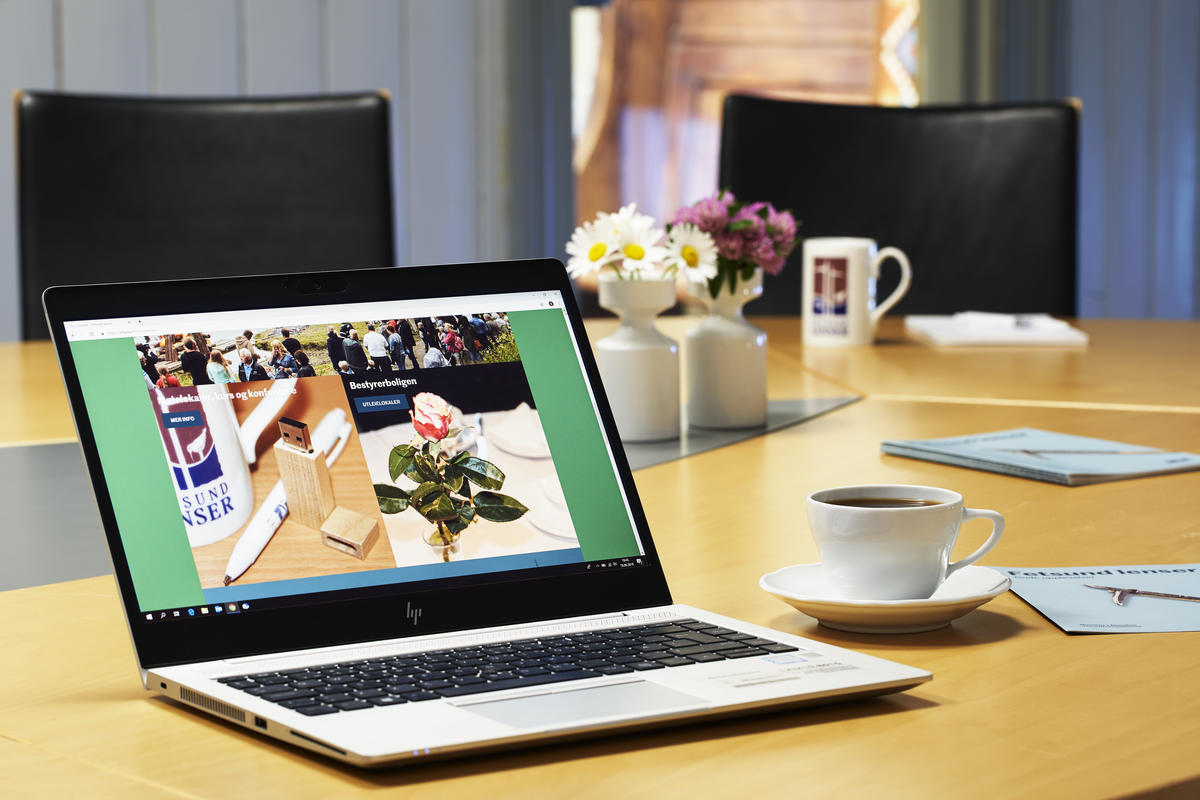 Bærbar pc i forgrunnen med kaffekopp og folder på høyre side på et konferansebord. Blomster og kaffekopper i bakgrunnen (Foto/Photo)