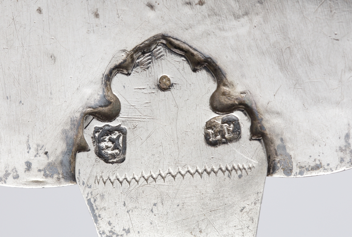 Tårtspade av silver. 
Bladet är utsträckt hjärtformat, med avrundad spets. Skaftet har en vinkelböj intill fästet, med en snäckformad avslutning längst bak. Ornamentsbården runt bladets översida är graverad och stämplad, i form av bågar, akantusrankor m.m. Skaftets översida har en liknande utsmyckning. 

Stämplar (mycket svårlästa, möjl. Lennart Montin, Stockholm, verksam 1726-1740) på skaftets infästning i bladet undertill, + åldermansranka.
Inskrivet i huvudbok 1936.