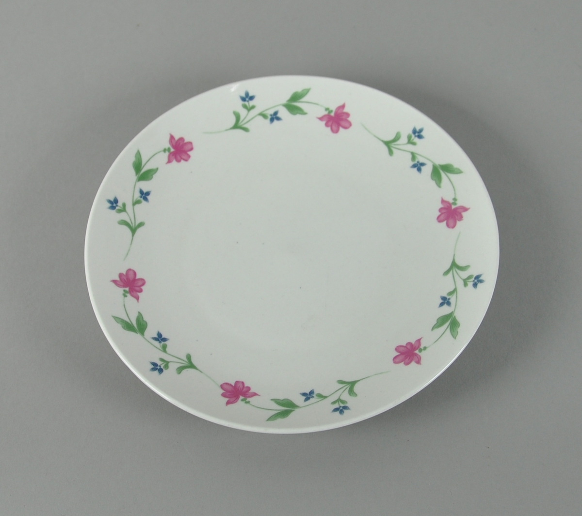 Hvit frokosttallerken av glassert keramikk, med blomsterbord av røde og blå blomster rundt kanten.