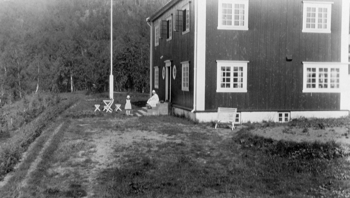 Skogforvalterboligen på Moen i Målselv i Troms. Fundamenteringsarbeidet for denne bygningen ble påbegynt i 1921, skogforvalter Harald Løkke og familien hans kunne flytte inn i ny embetsbolig våren 1923, året etter at han tiltrådte dette embetet.  Dette fotografiet skal være tatt i 1926, etter at bygningen var malt utvendig. Det dreier seg om en forholdsvis stor toetasjes bygning, antakelig en reisverkskonstruksjon som utvendig var kledd med vekselpanel. Vinduene er smårutete - hovedsakelig to-rams, men tre-rams i et rom til høyre på bildet, muligens kjøkkenet. På begge sider av hovedinngangsdøra på langveggen var det små, runde vinduer, som antakelig skulle slippe lys inn i en bakenforliggende gang. Framfor denne døra var det ei støpt trapp, der det satt ei kvitkledd kvinne. Framfor henne sto det en kjolekledd jentunge. Bakenfor barnet sto det et bord med krakker og ei flaggstang. Framfor huset var det plantet en hekk. 

Skogforvalter Harald Løkke og kona Juliane hadde bodd i Troms i fire år da dette fotografiet ble tatt. Da de kom ble de antakelig innkvartert på garden Olsborg som den statlige skogetaten hadde kjøpt av en tidligere skogforvalter, Anders Sakshaug, i 1916. Sakshaug hadde altså holdt seg sjøl med bolig. Bygginga av det store og solide huset på bildet var antakelig et ledd i et program for å skaffe skogforvalterne i alle regioner gode og tidsmessige boliger. Løkke ble i skogforvalterstillingen i Troms til 1935.