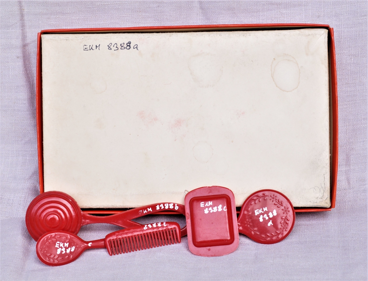 Lises dukkesett i en eske (a) som inneholder rangle (b), såpeskål (c), speil (d), børste (e), og kam (f) av rød plast.