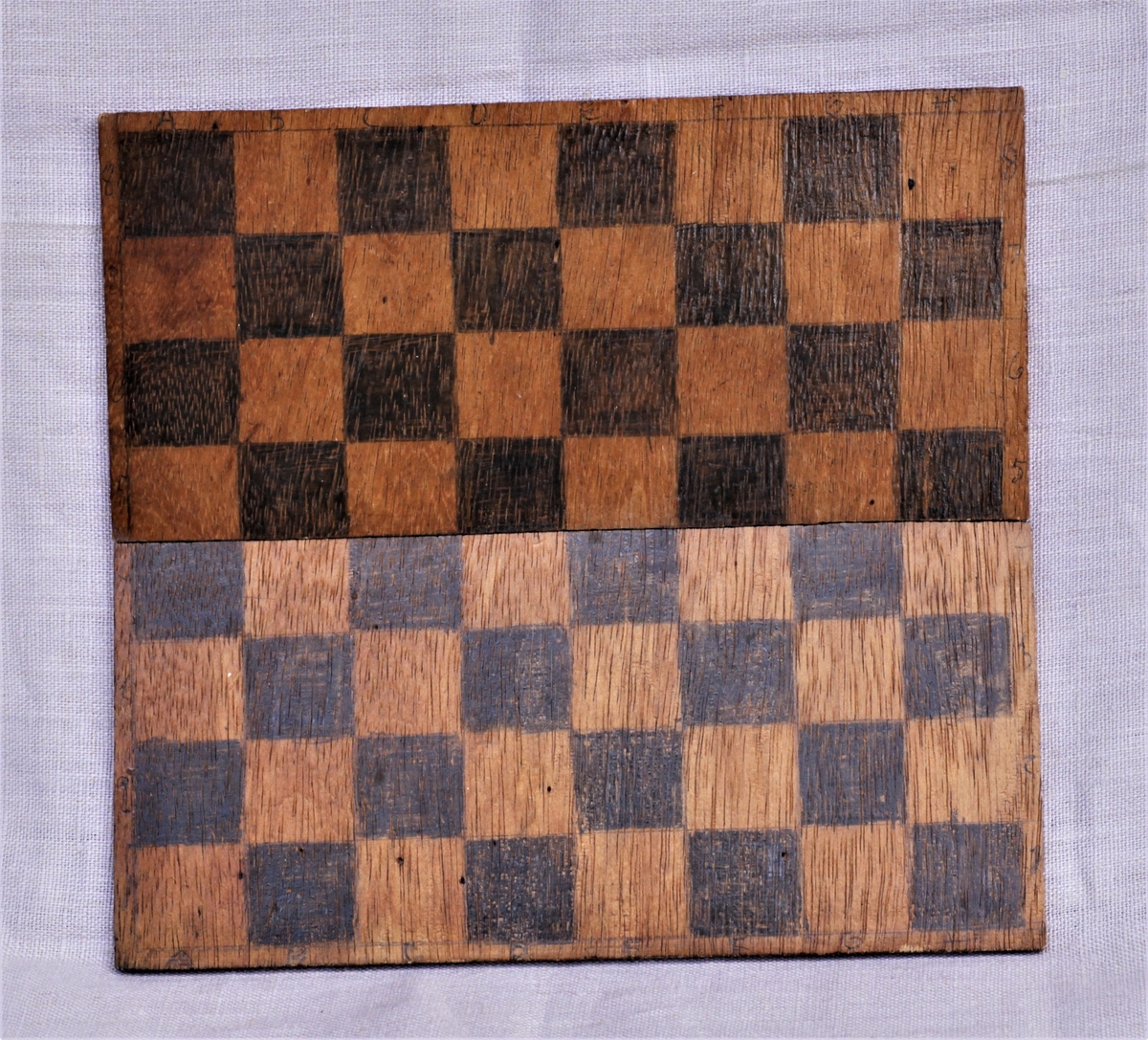 Hjemmelaget sjakkbrett. Utskjært treplate i to deler, festet med tape på baksiden. Dette er gjort slik at den kan legges sammen. Tegnede felter, tall og bokstaver.