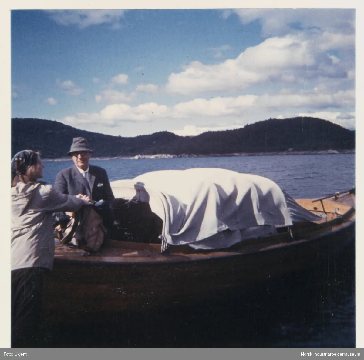 James Coward står i snekke med kalesje på Møsvatn. Unni Coward står på land og legger veske på båten