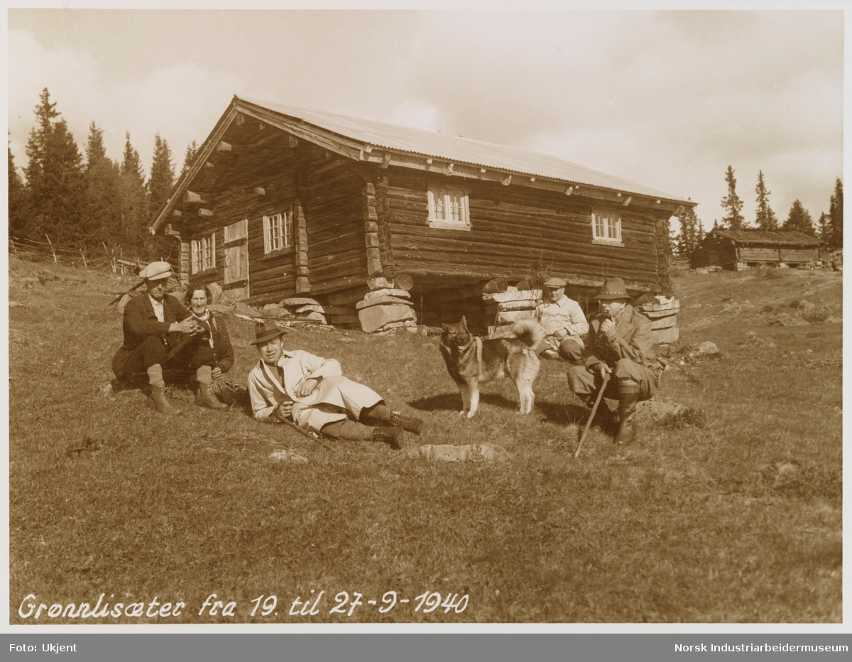 Mennesker sitter og ligger på gress på Grønnli seter. I bakgrunn sees laftet hytte og gårdshus