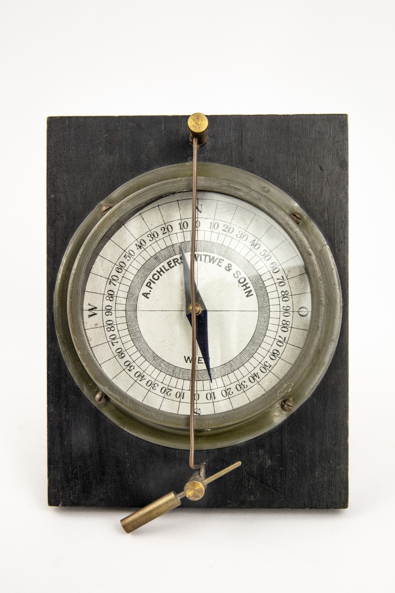 En kompass med graderad platta under en glasskiva. Kompassen är fastsatt på en bottenplatta i trä och rakt över hela plattan löper en metallsträng.