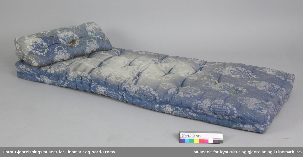 Den blå madrass har en tilhørende kilepute. de er laget av tettvevd stoff med blå-hvit mønster. Fyllet holdes på plass av snøringer av naturhvite og grønne bomullstråder.  