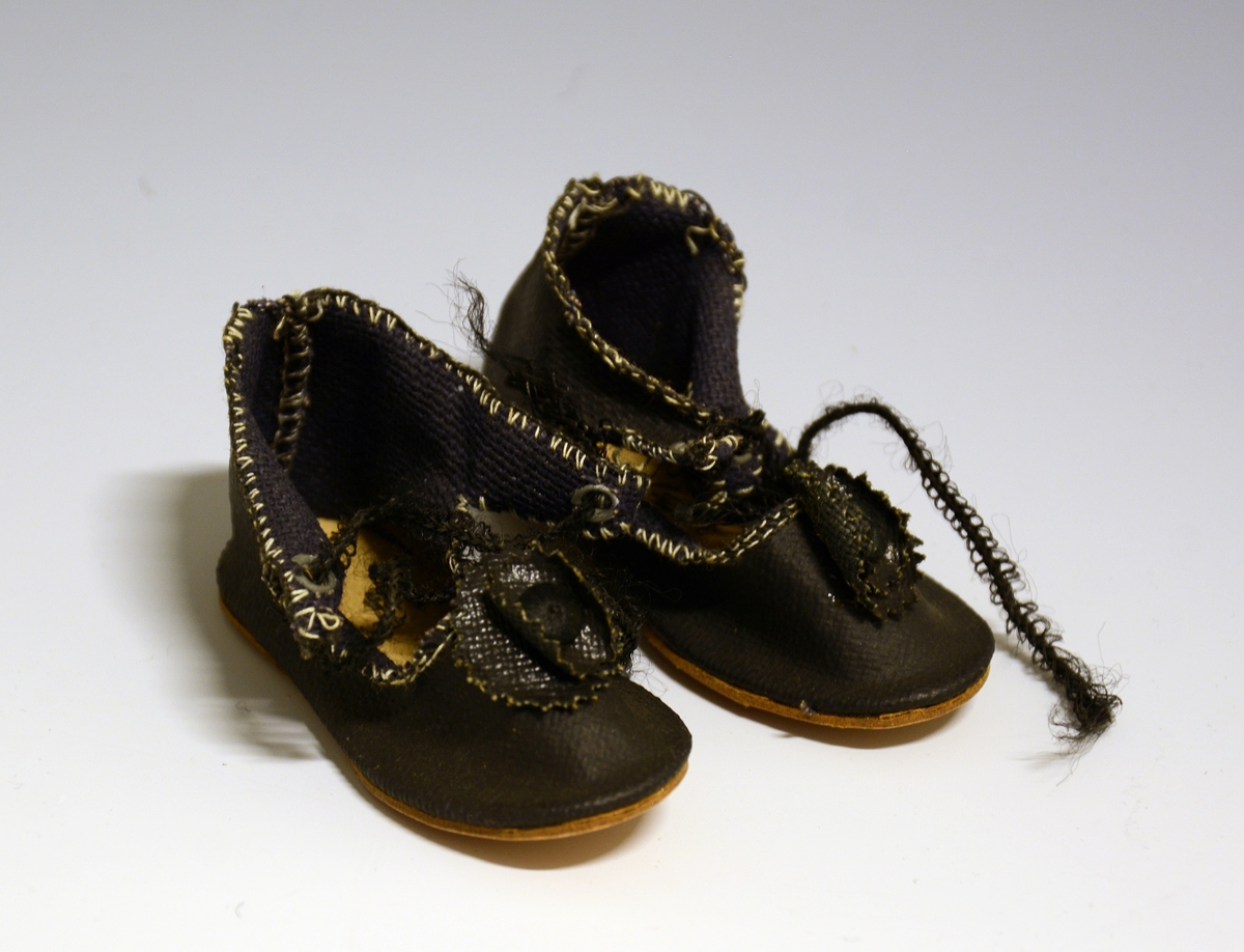 Et par pensko til dukke i sort tekstil. Åpent nederst på snørestykket, med lisser og dusk. Lys brun lærsåle.
