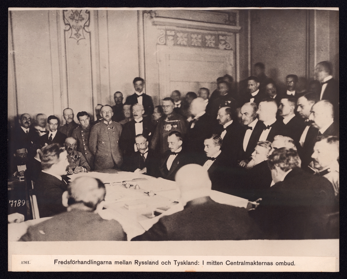 Bilden föreställer fredsförhandlingarna mellan Ryssland och Tyskland. Man ser ett fler tal finklädda herrar som sitter runt ett bord. Det omgivande rumet är fyllt med fler herrar med civil beklädnad och uniformer.