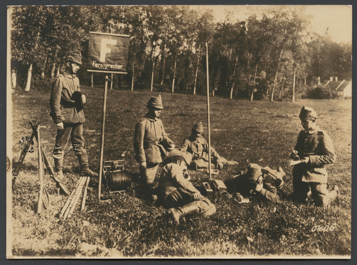 Bilden visar en grupp soldater som ligger på en gräsmatta där de har upprättat en mobil telefonstation. Skyltet "Fernsprecher" hänvisar till att det finns en telefon. En av männen ligger på mage och prata i hörluren medan han skriver. På vänster sidan ser man en kabeltrumma och tre gevär.