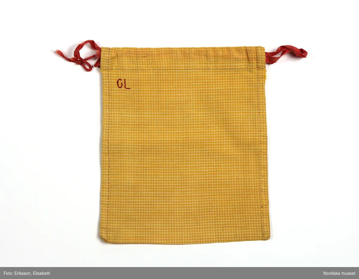 Påse sydd av gult tyg med tunna vita rutor. Handsydd med röd tråd. Dragsko och röda bomullsband. Monogram. 
/Fiffi Myrström, 2014-02-24