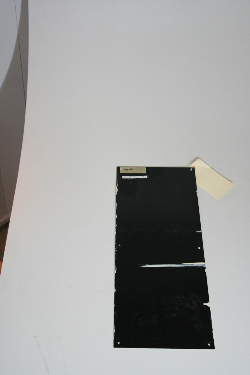 Rektangulørt svart skilt med hvit skrift i en serie av 4 skilt
6 hull for skruer.
Skader i kanten rundt skiltet.
Skadet bakside. En vertikal stripe midt på skiltet er oppskrapet slik at den sorte fargen er borte. Hvit farge kommer frem.